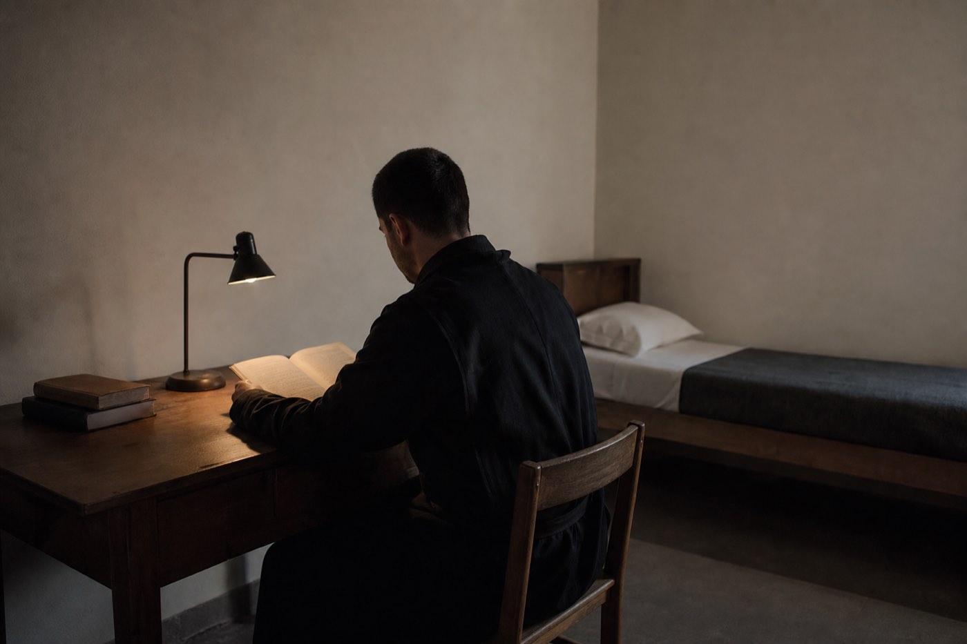 A solitary figure at a desk lit by a single lamp, turned away from the room behind them.